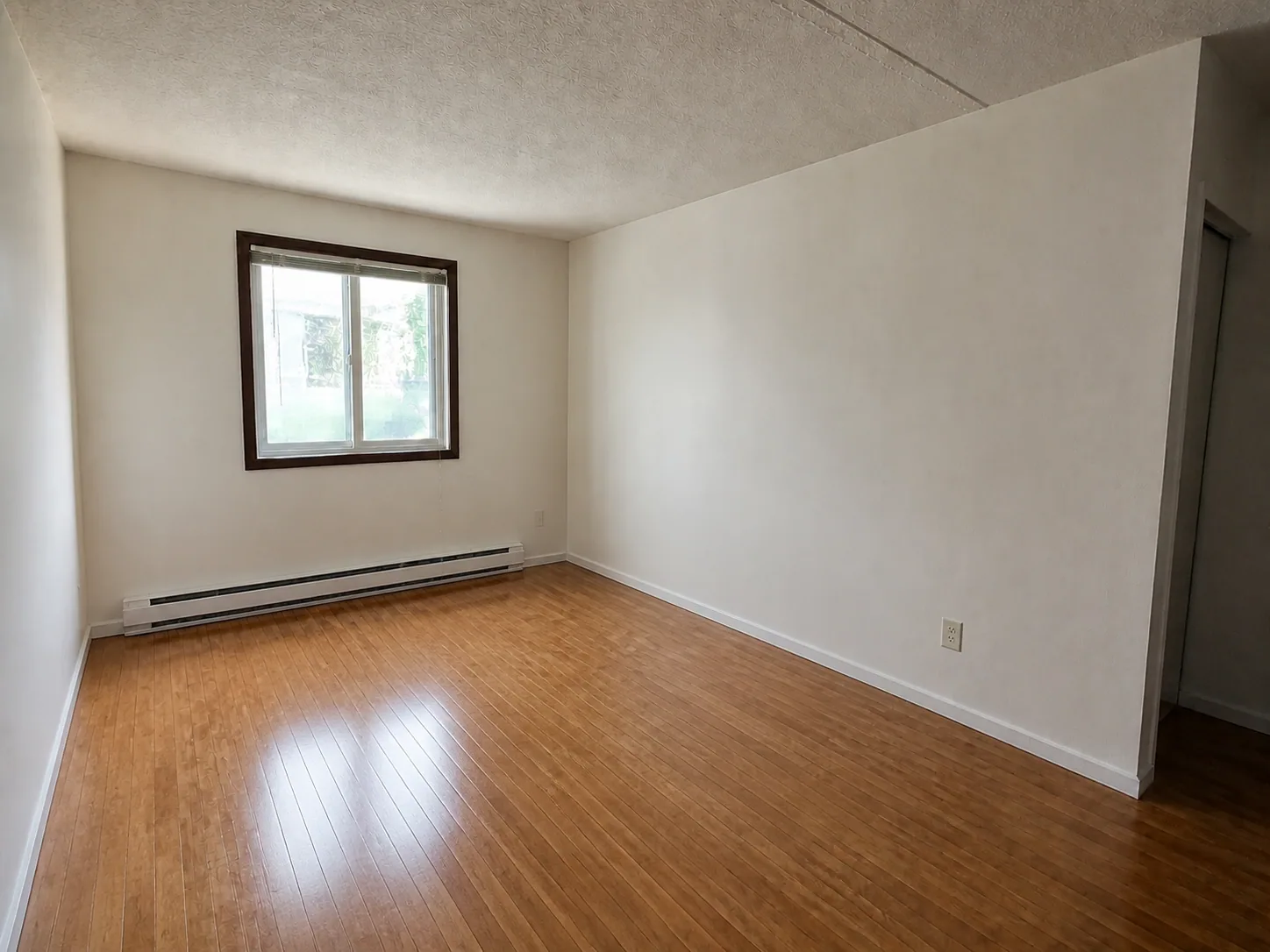 Empty apartment room with a wood floor, one window, and a baseboard heater